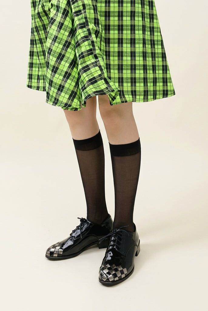 A person wearing a bright green and black plaid skirt, black knee-high socks, and shiny black-and-white patterned lace-up shoes, standing on white pebbles outdoors.