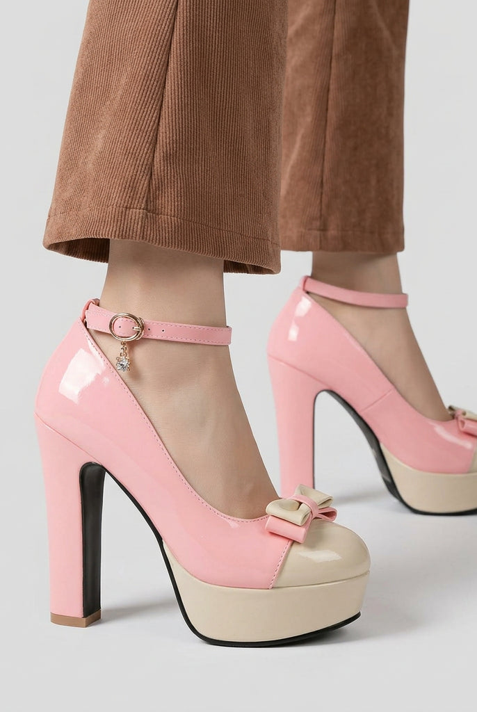 A pair of women's high-heeled platform sandals in light pink, shown being worn with brown pants on a wooden floor. The shoes feature a chunky block heel, a cream-colored platform sole, an ankle strap with a gold buckle, and a small bow detail on the toe.
