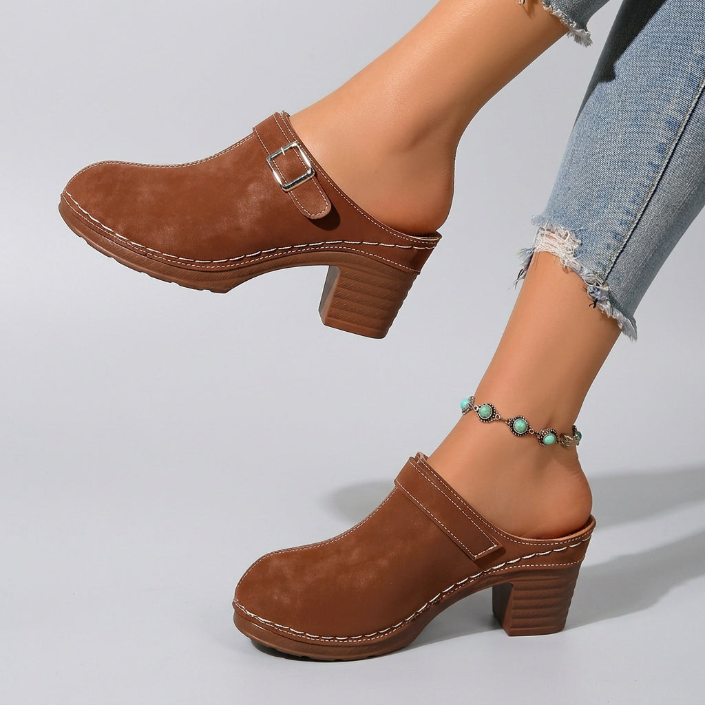A pair of women's brown suede clogs with block heels and buckle straps, shown being worn outdoors on a tiled surface with a stone wall background.
